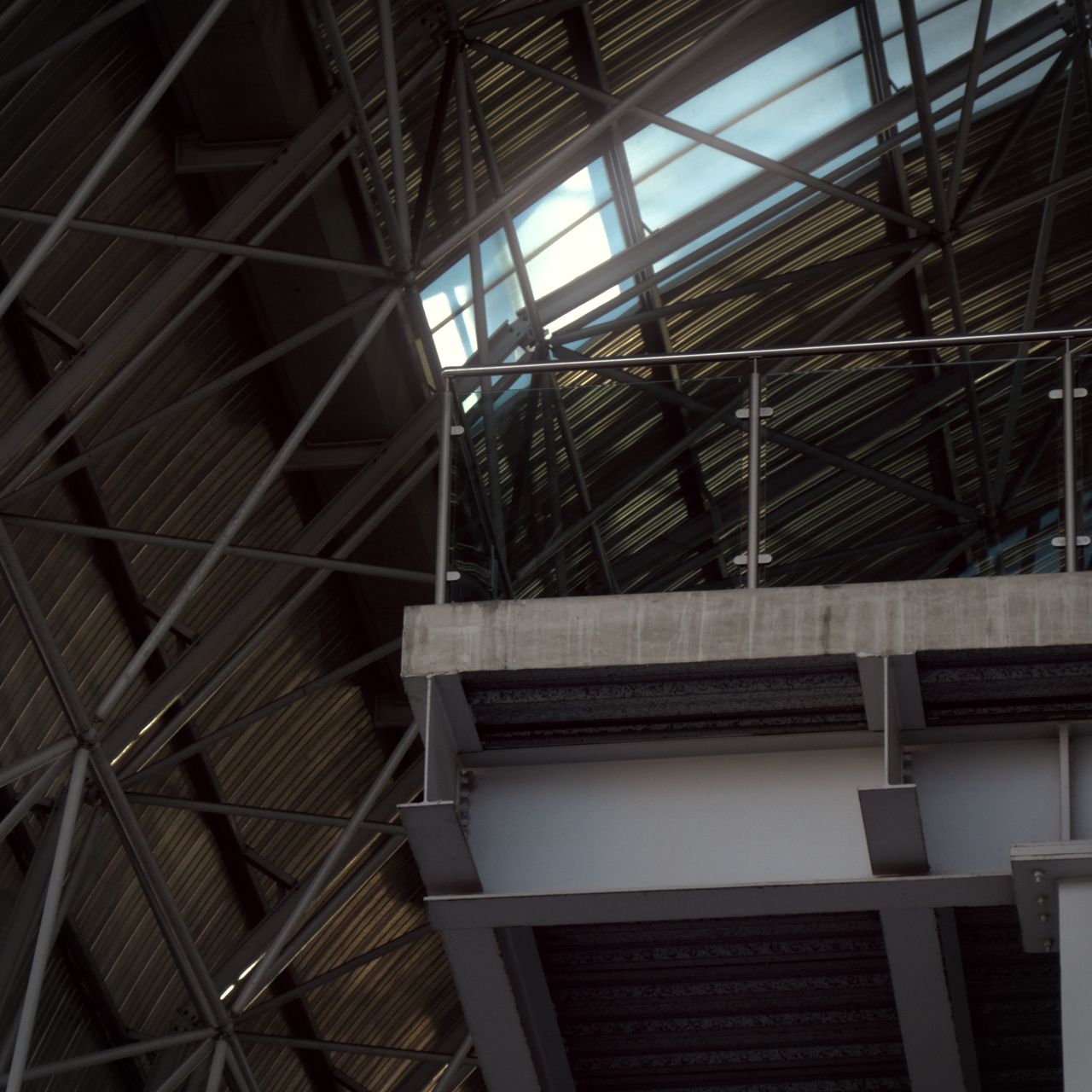 A floating platform edge with a glass railing sticks out beneath a steel-framed ceiling. Sunlight shines through the roof above, highlighting the suspended structure.