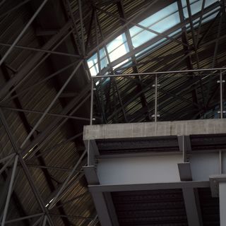 A floating platform edge with a glass railing sticks out beneath a steel-framed ceiling. Sunlight shines through the roof above, highlighting the suspended structure.